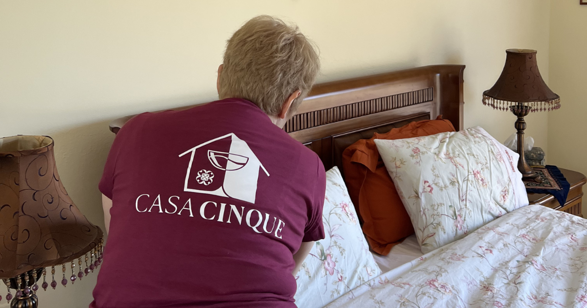 A person wearing a Casa Cinque shirt prepares a bed in a guest room.